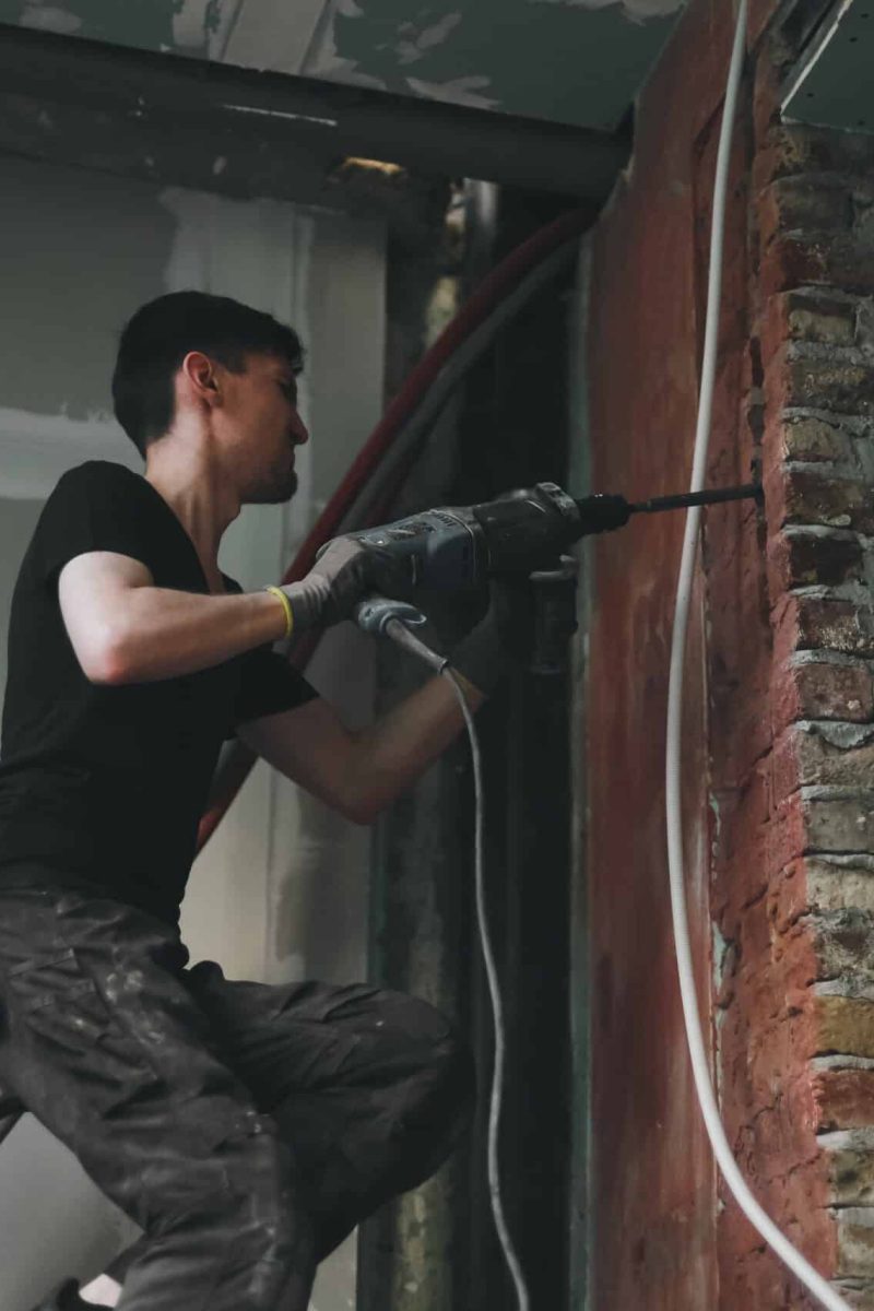 A young man breaks a wall with a perforator. A young Caucasian man recognizable in a black uniform emotionally breaks a hole in a brick wall with a puncher with a large screwdriver while sitting on a stepladder,close-up bottom view with selective focus. The concept of home renovation, construction work.