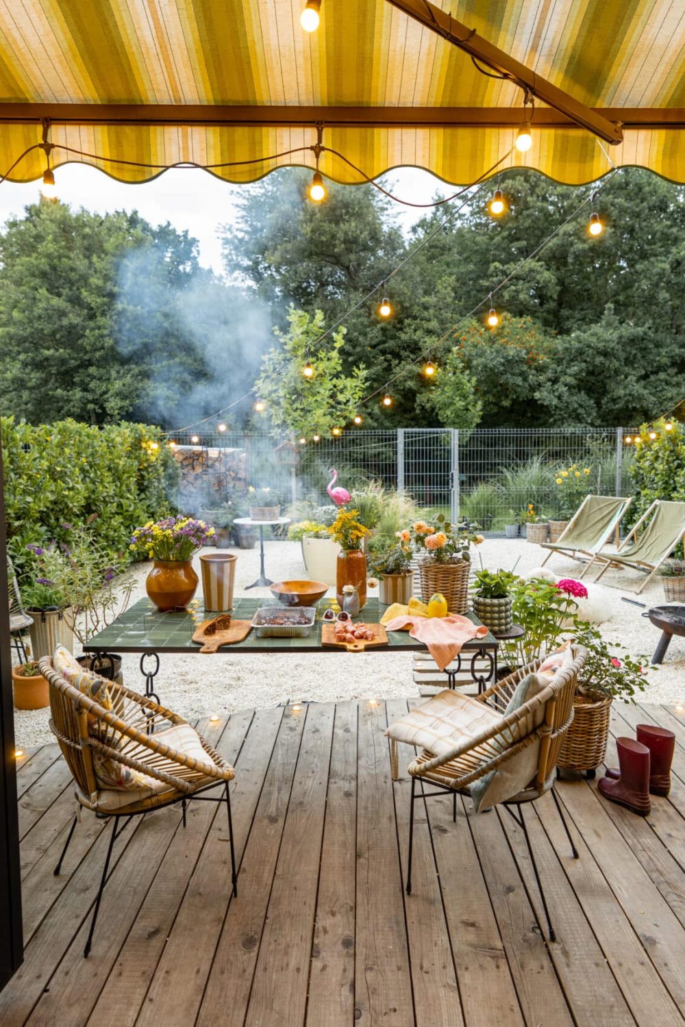Atmospheric and cozy garden with dining place on terrace at dusk. Cooking food on disposable grill on beautiful table decorated with flowers. Wide view from the house