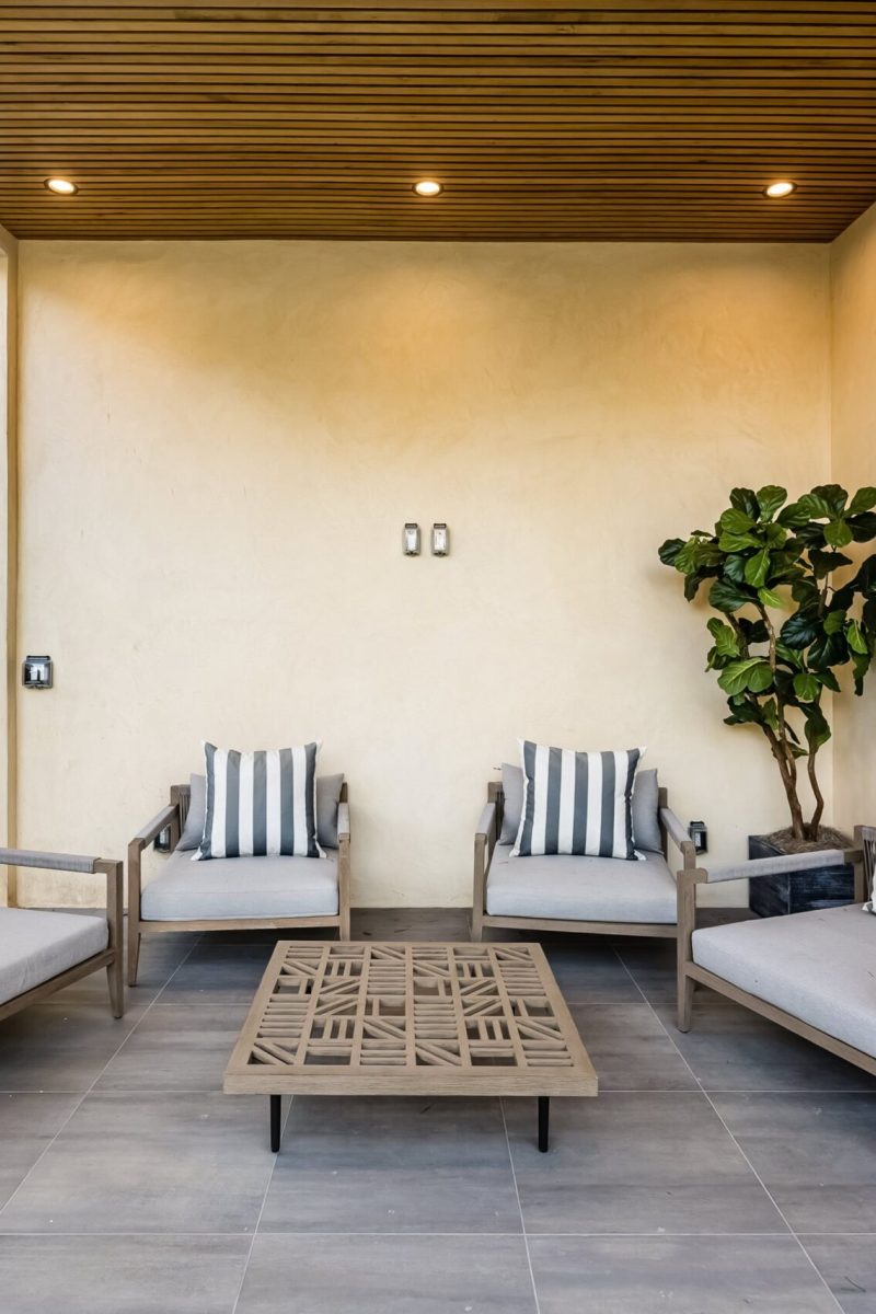 Modern outdoor patio with wooden ceiling, cushioned chairs, striped pillows, and a coffee table, surrounded by greenery.