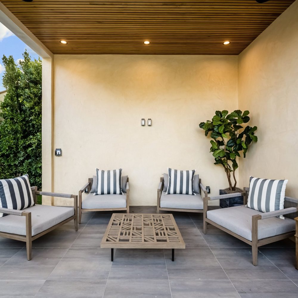 Modern outdoor patio with wooden ceiling, cushioned chairs, striped pillows, and a coffee table, surrounded by greenery.